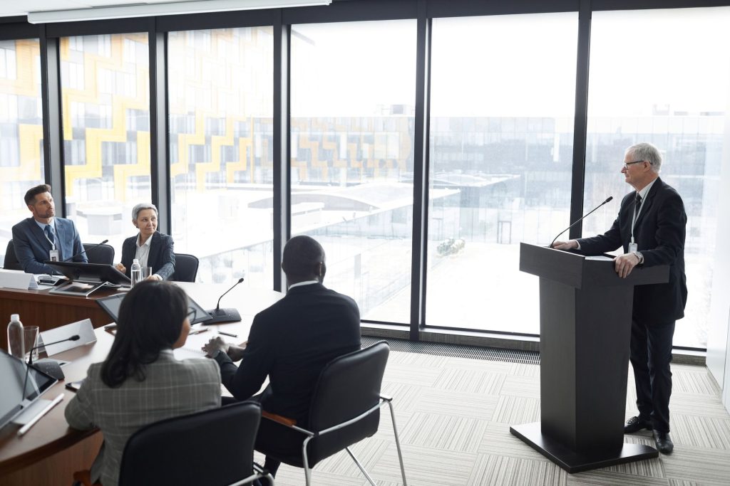 A senior speaker addresses four colleagues in a formal conference room with microphones and individual meeting consoles.